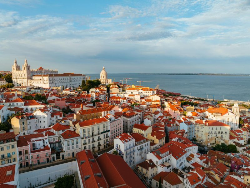 the-lakhani-group-global-lisbon-website Aerial view of the historic Alfama district with red-roofed buildings in Lisbon, Portugal, overlooking the Tagus River.